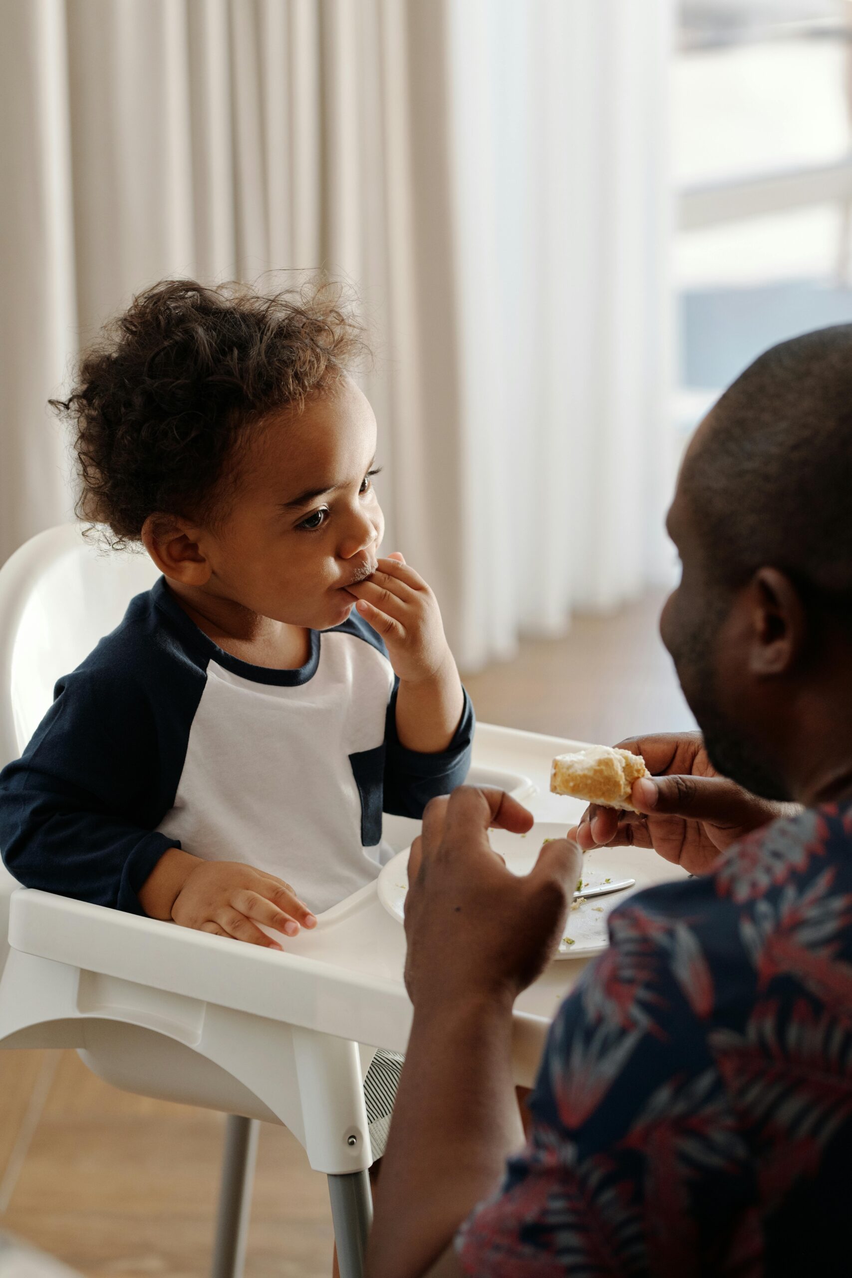 A joyful family moment of a father feeding his baby in a high chair indoors.